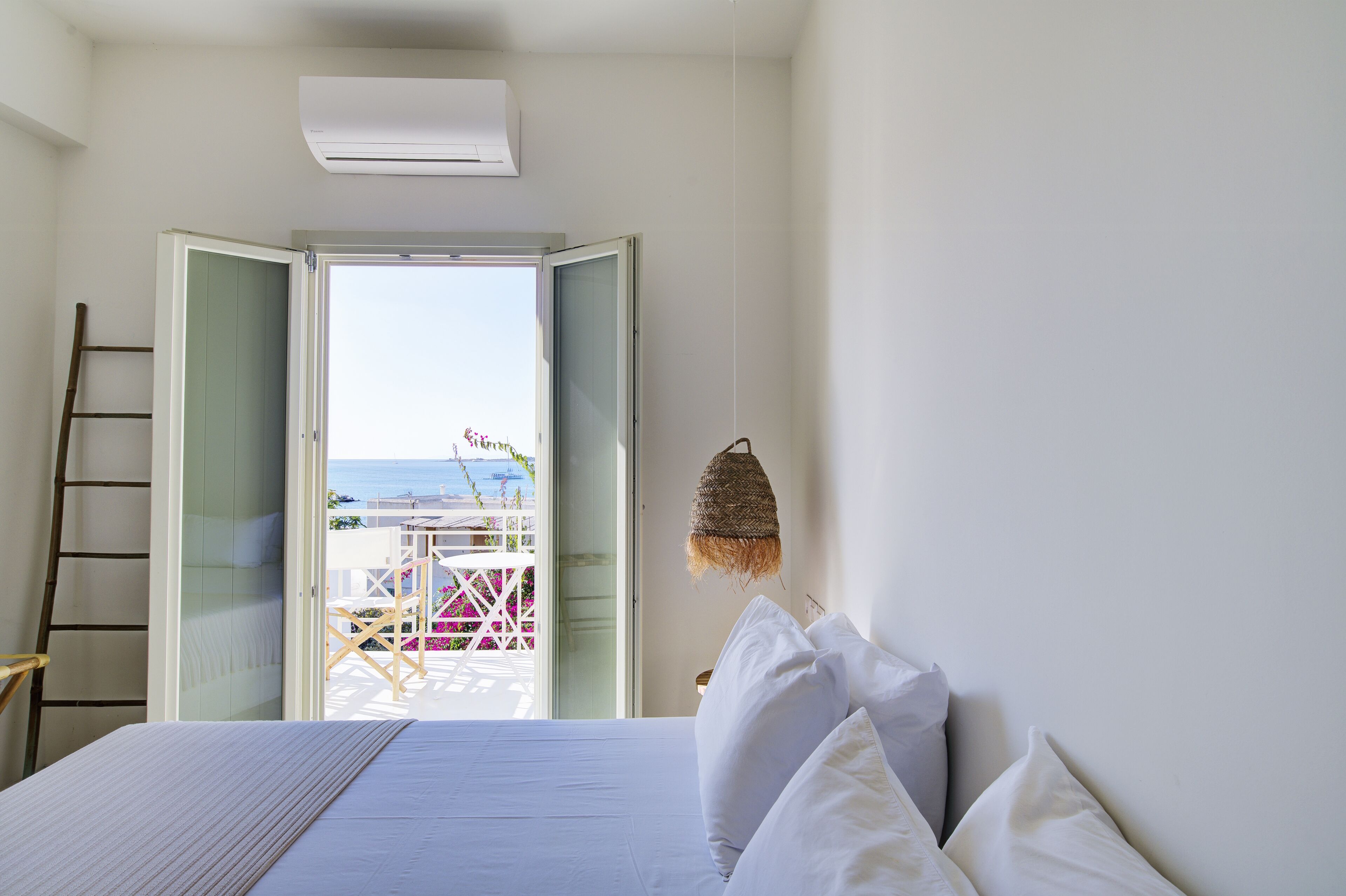 A bedroom with a bed, white pillows, a hanging basket, and an open door leading to a balcony with a view of the sea.
