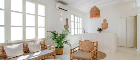 A bright, airy room with a wooden sofa, a chair, and a small table. There is a potted plant and a hanging light fixture.