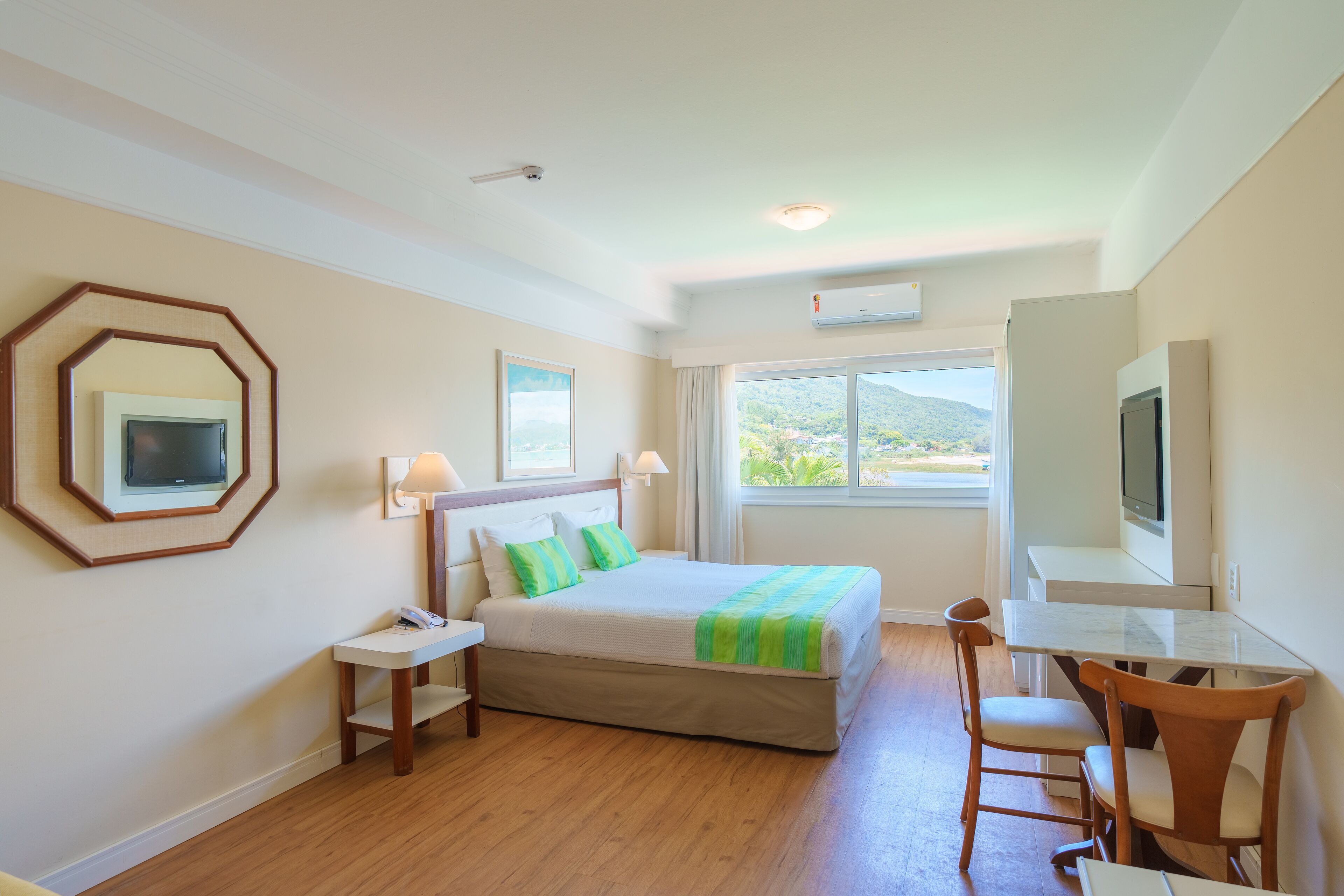 Quarto de hotel com cama, escrivaninha, cadeira, televisão e janela com vista para o verde.
