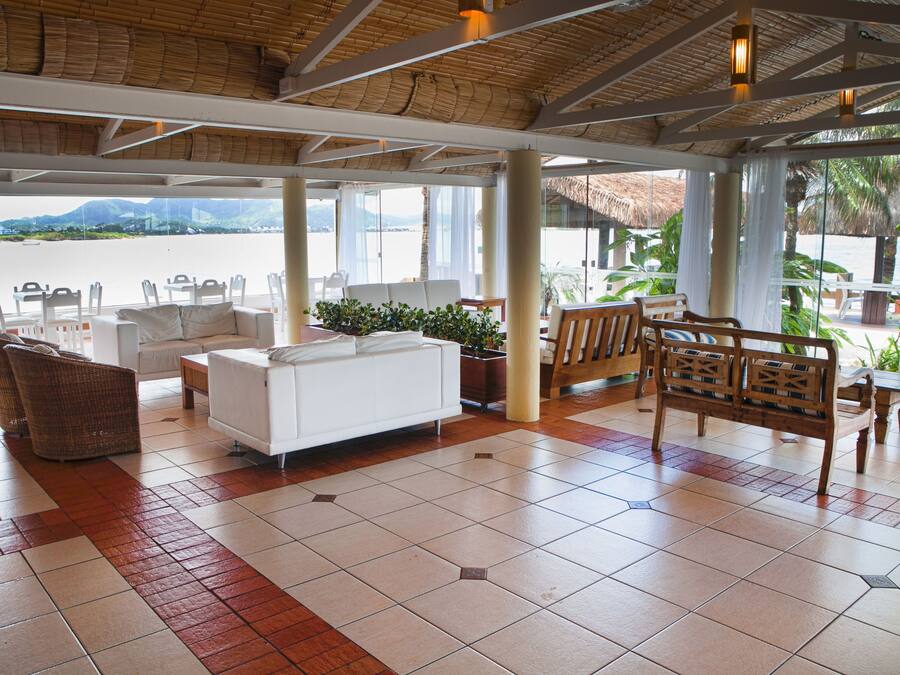 Amplio espacio interior con un sofá blanco, sillas de mimbre y un banco de madera. Grandes ventanales ofrecen vistas al mar y a las palmeras.