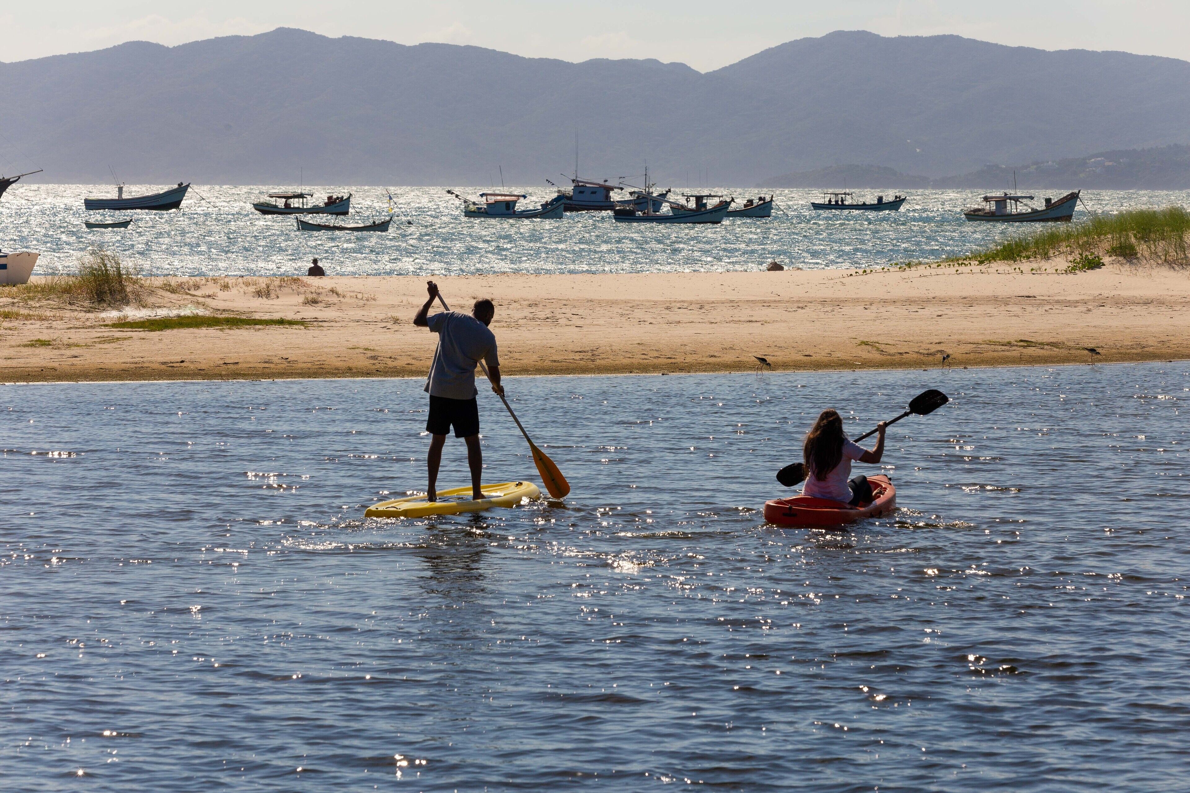 Une plage où l’on peut faire du paddle et du kayak, avec des bateaux à l’arrière-plan et des montagnes à l’horizon.