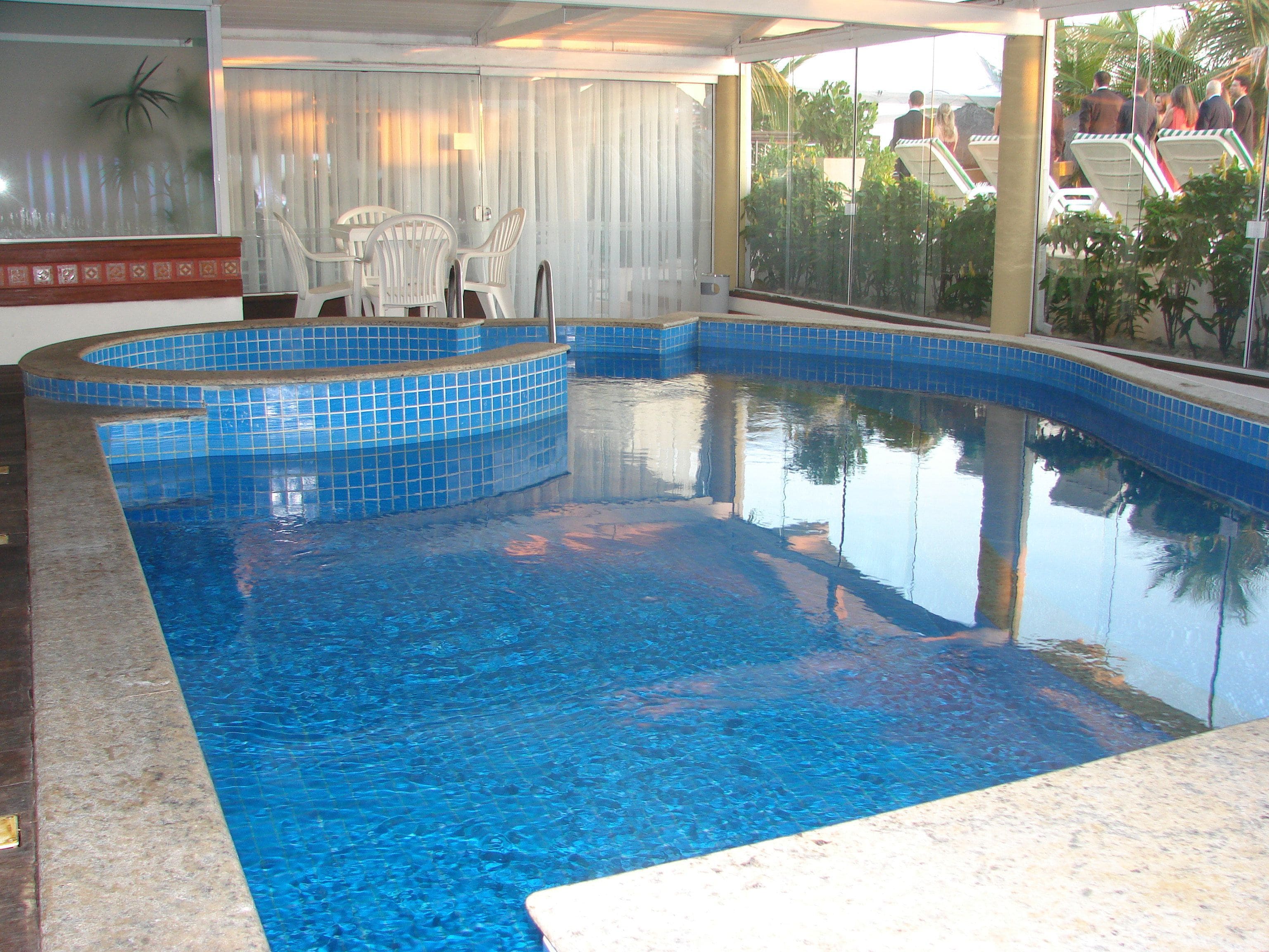 Una piscina con azulejos azules, una zona de estar con sillas y vistas a palmeras y un edificio.