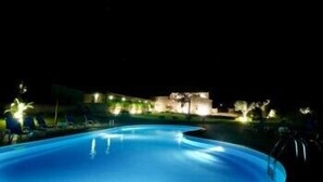 A nighttime swimming pool with a curved edge, surrounded by a landscaped area with trees and a building in the background. - Torre Dantona (Modica)