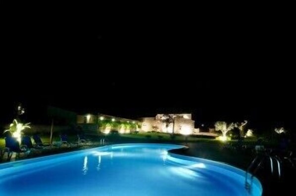 A nighttime swimming pool with a curved edge, surrounded by a landscaped area with trees and a building in the background. - Torre Dantona (Modica)
