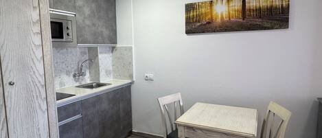 A compact kitchen with a microwave, sink, and cabinets. There is a dining table with chairs and a painting on the wall.