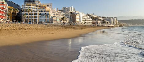 A beachfront hotel with multiple stories, a sandy beach, and a clear sky.