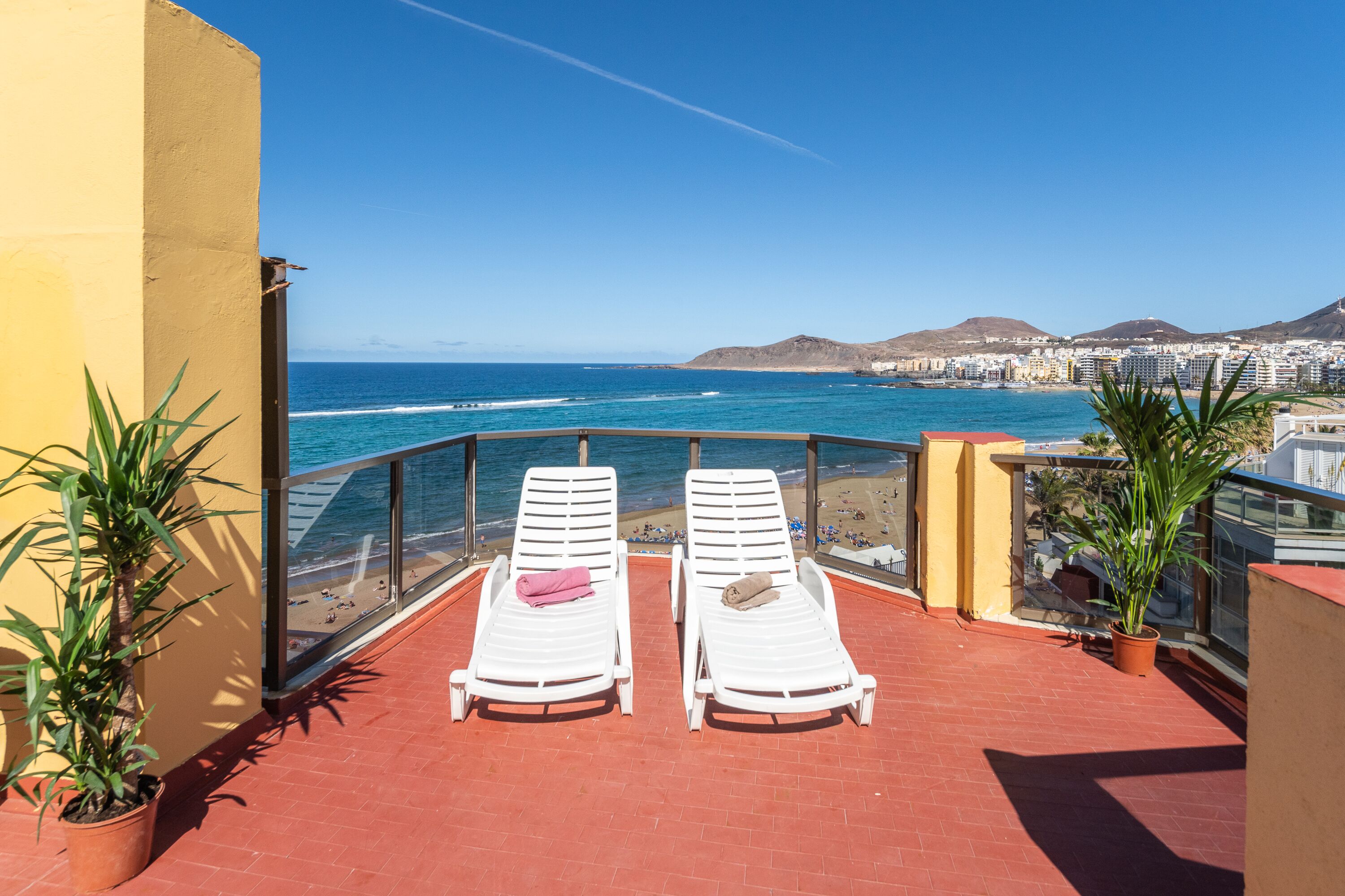 A balcony with two white sun loungers, a potted plant, and a view of the ocean and cityscape.
