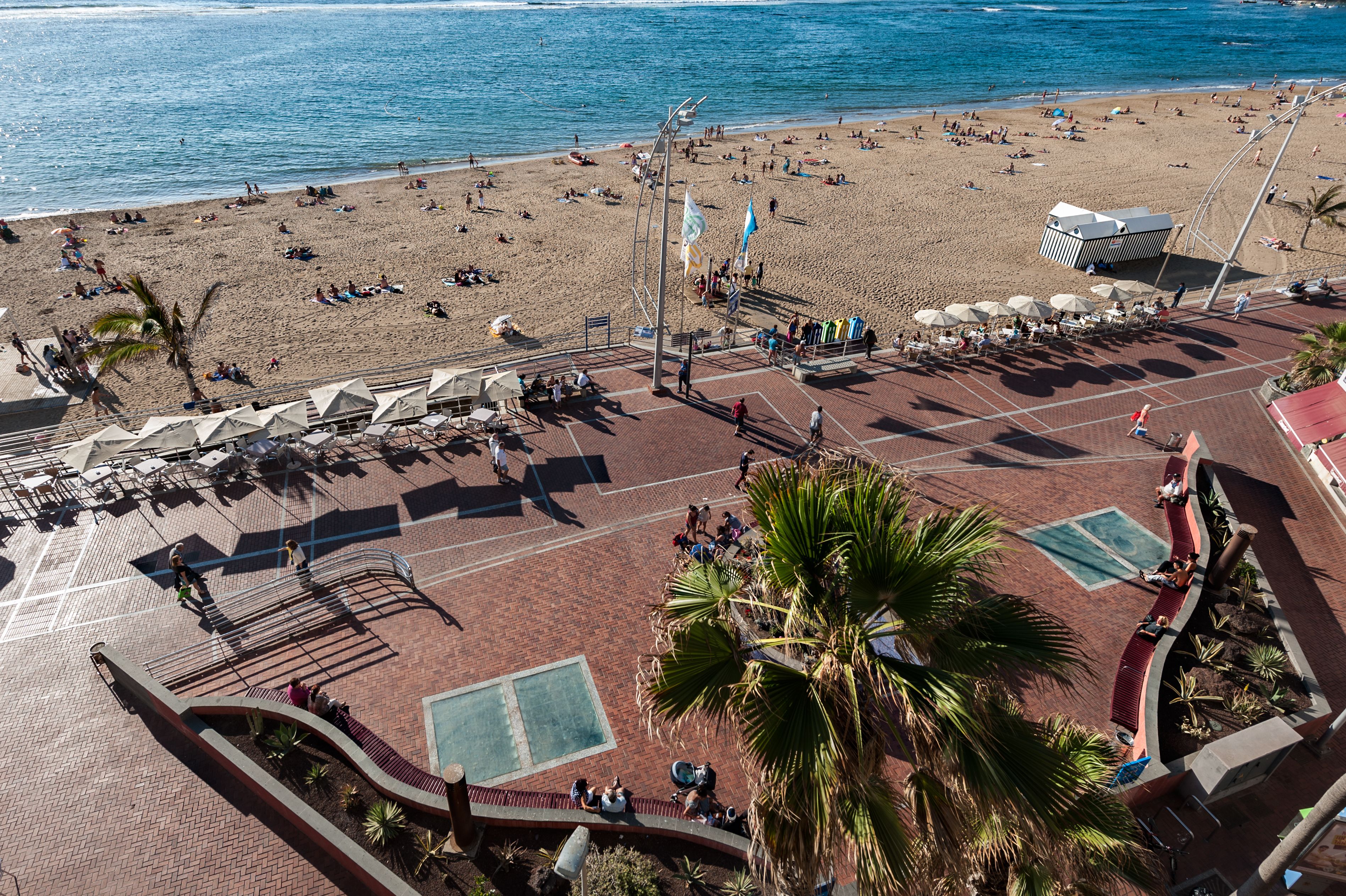 Een overzicht vanuit de lucht van een strand met een verharde promenade, palmbomen en strandparasols.