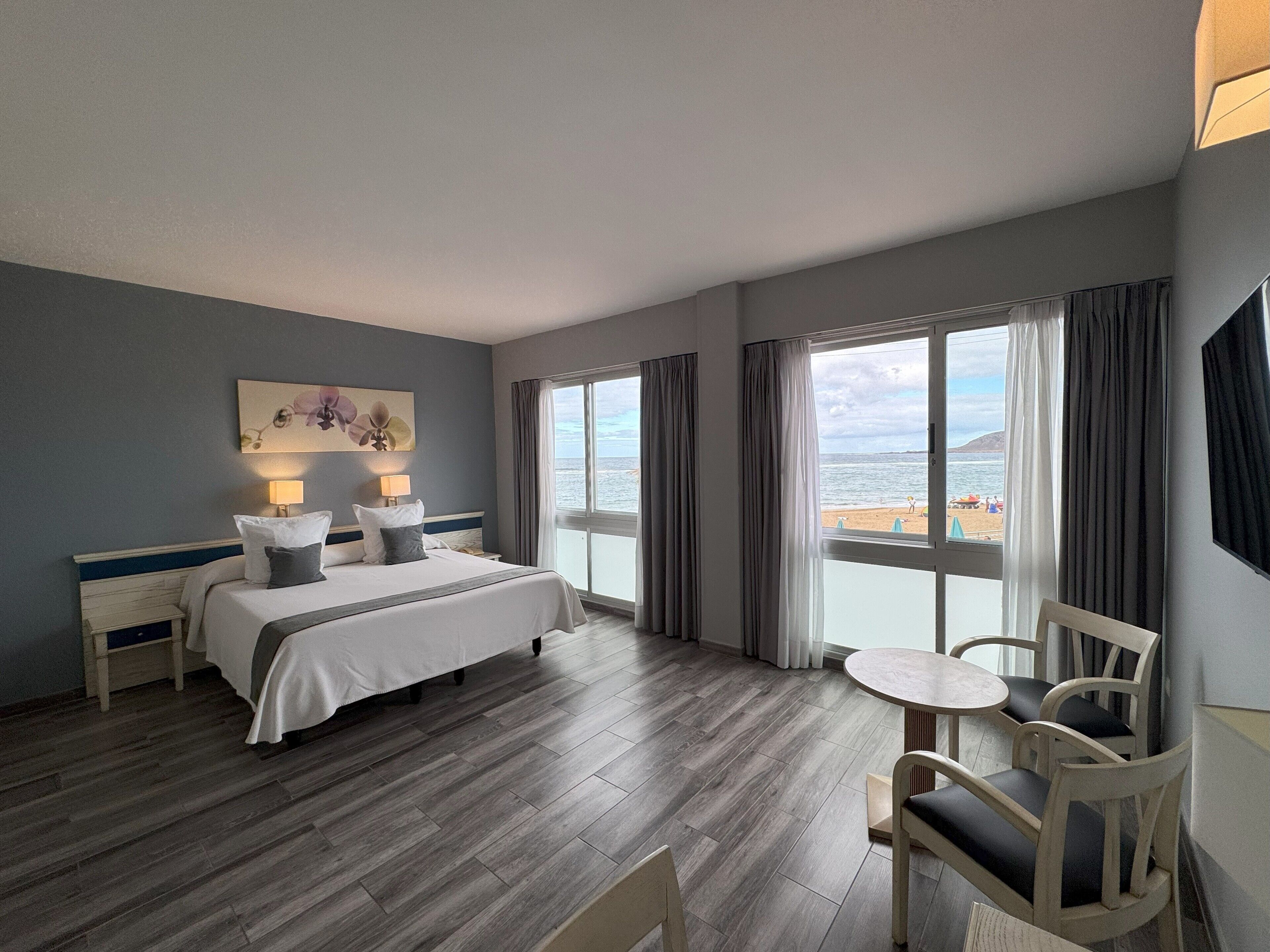 Habitación de hotel con cama, una mesa y sillas, vista a la playa y un cuadro en la pared.