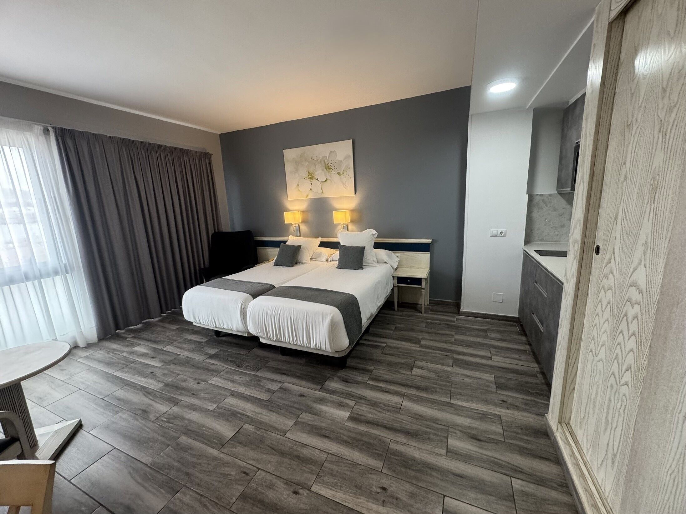 A hotel room with a double bed, bedside tables with lamps, a small desk, a chair, and a large window with curtains.