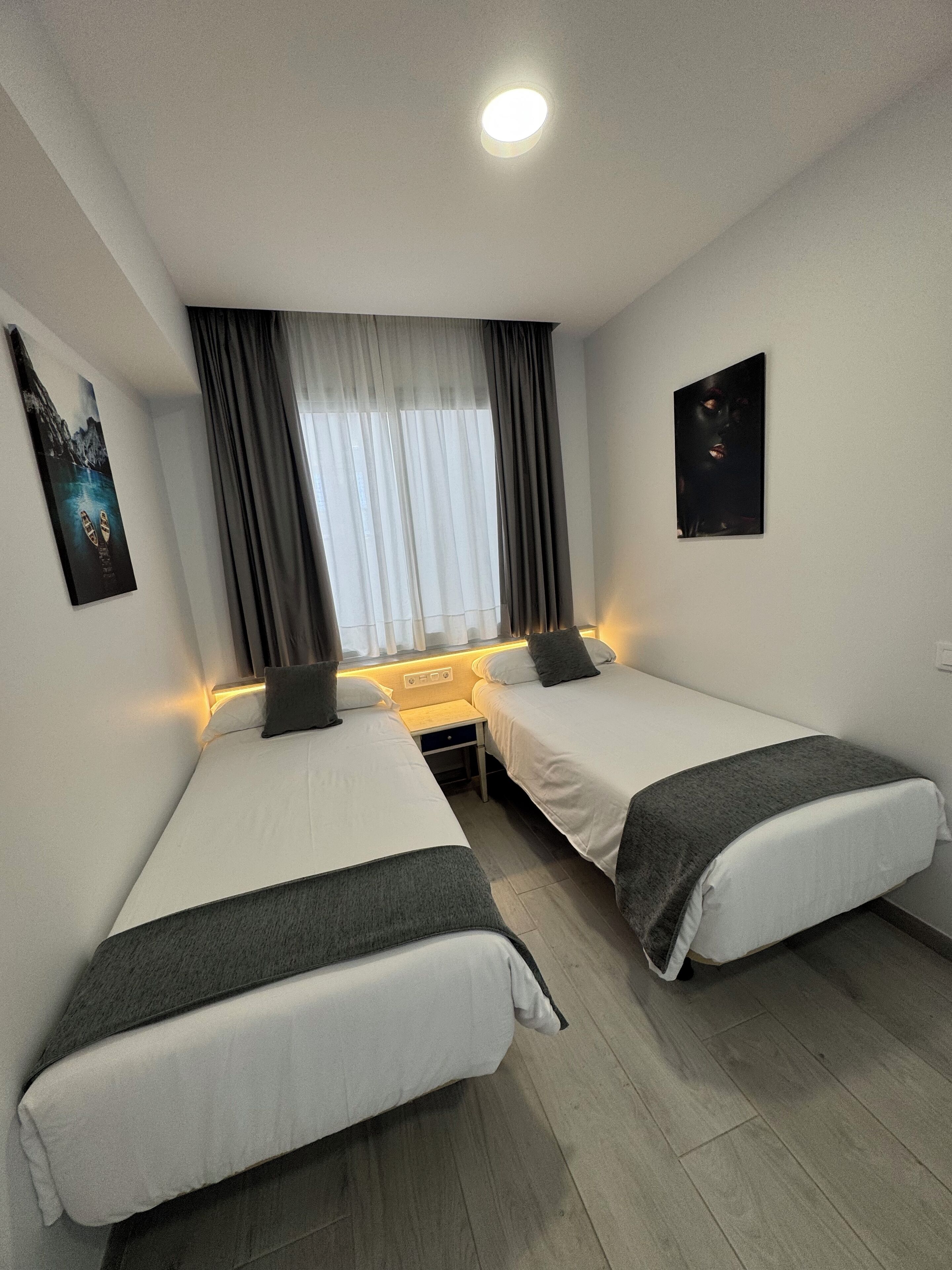 A hotel room with two single beds, a nightstand, a lamp, a window with curtains, and artwork on the walls.