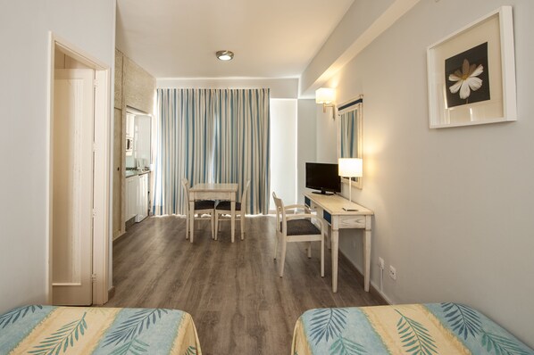 Une chambre d’hôtel avec un plancher en bois, un bureau avec une chaise, une télévision, un lit recouvert d’une couvre-lit à motifs de feuilles, une porte donnant sur une salle de bain et un rideau à rayures.