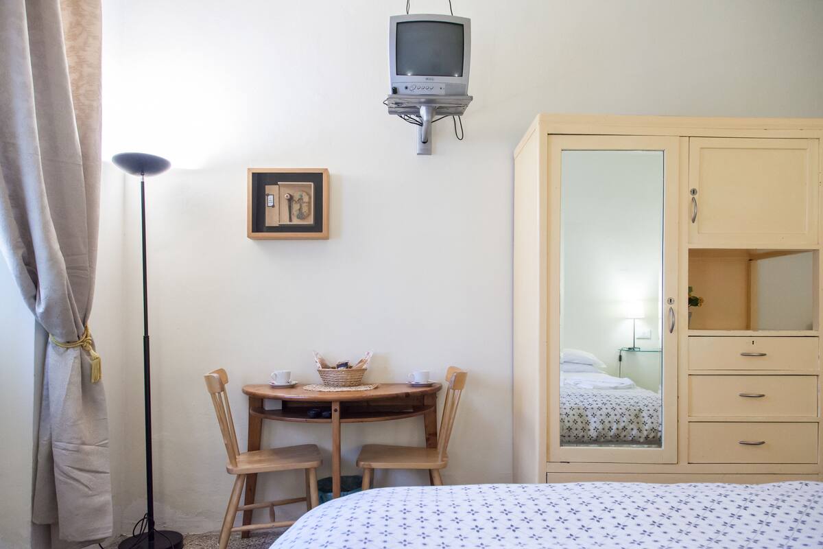 A room with a bed, a wooden table with chairs, a television mounted on the wall, a wardrobe, and a framed picture on the wall.