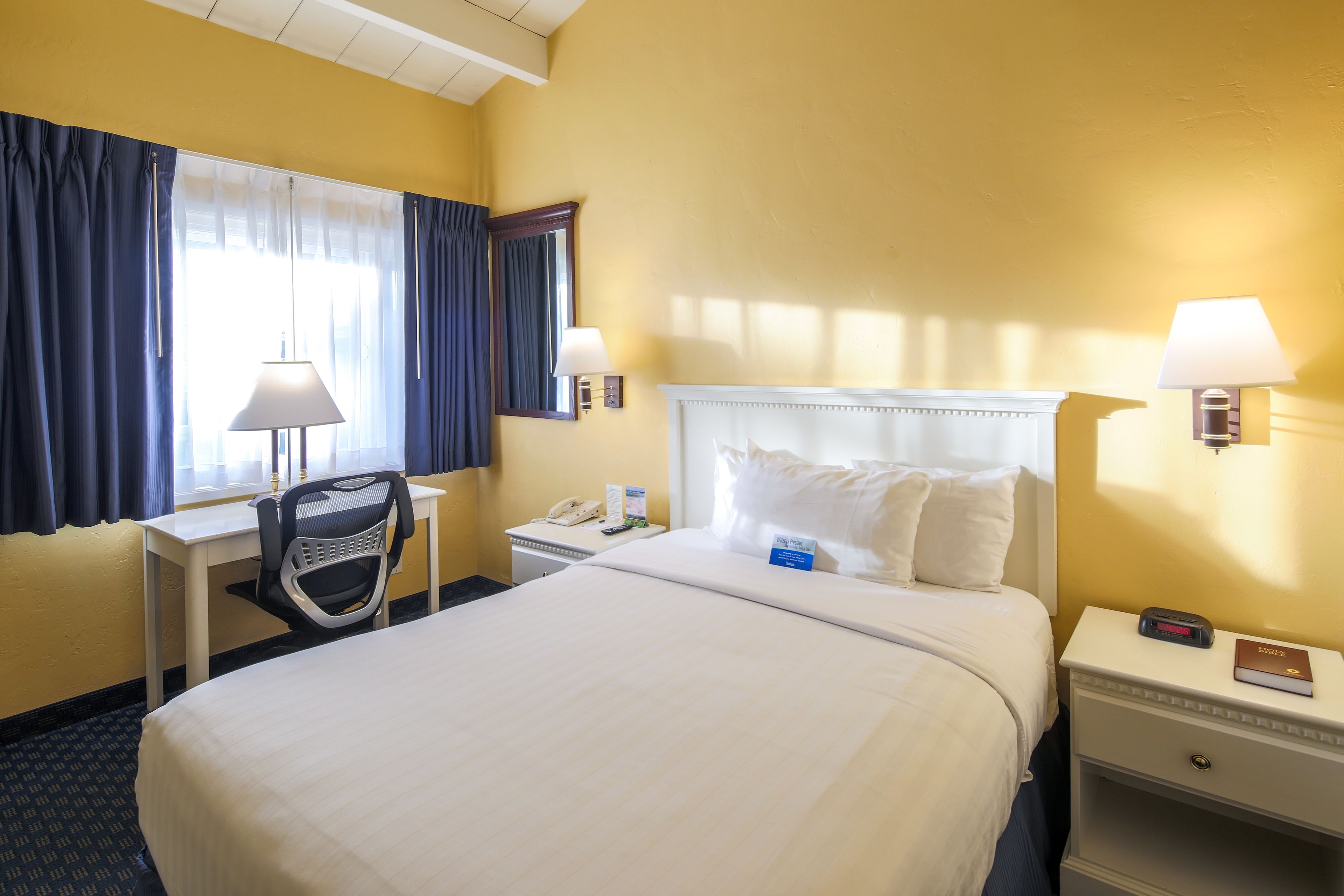 Une chambre d’hôtel avec un grand lit, un bureau, une chaise, une table de chevet, une lampe et une fenêtre avec des rideaux.