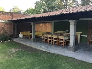 Un patio cubierto con pared de ladrillo, pared verde y techo de baldosas. Hay sillas de madera y un banco bajo el techo.
