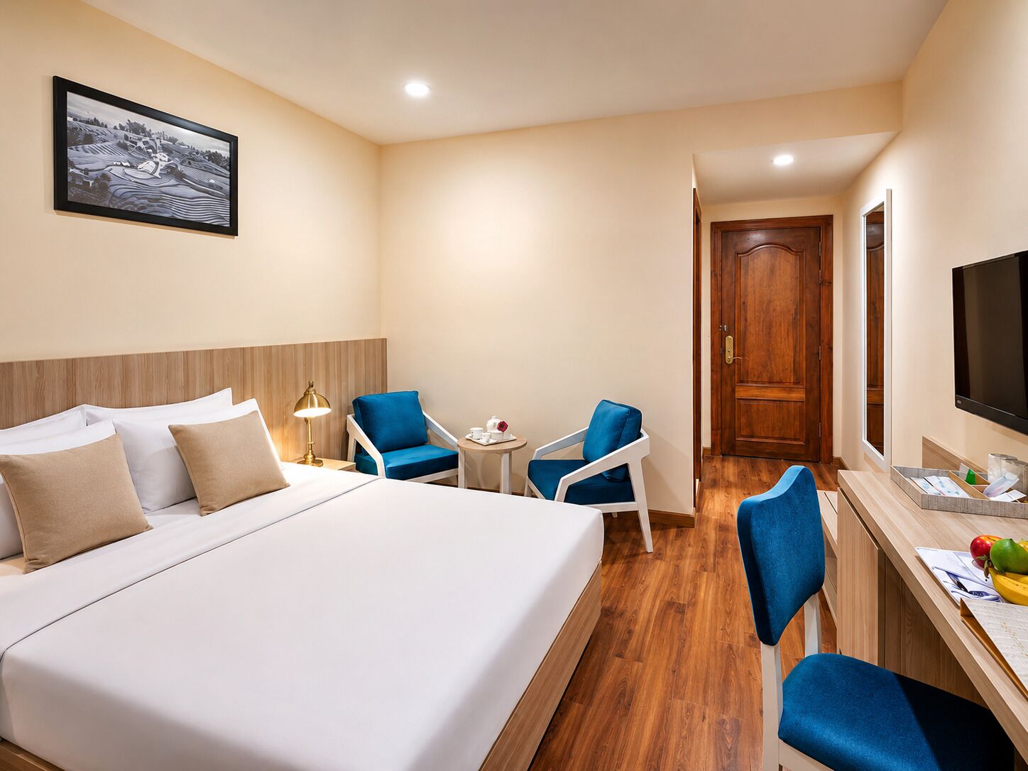 Une chambre d’hôtel avec un lit, deux chaises bleues, une petite table avec une lampe, un téléviseur et une porte en bois.