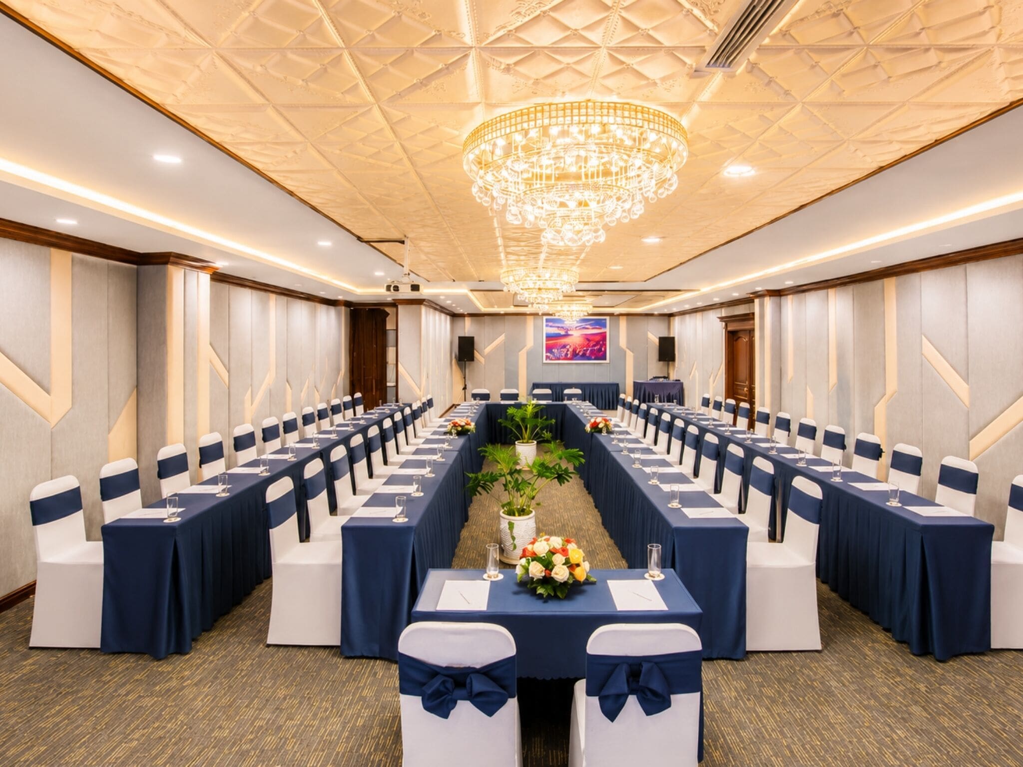 Un salón de conferencias con mesas largas, sillas, una araña y una pantalla grande.
