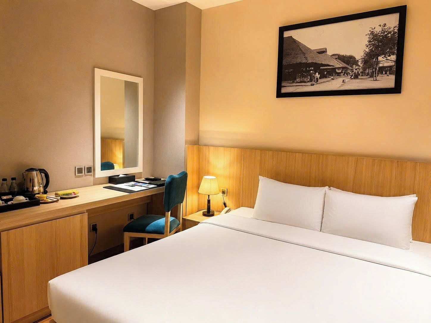 A hotel room with a bed, a desk with a chair, a lamp, a mirror, and a framed picture on the wall.