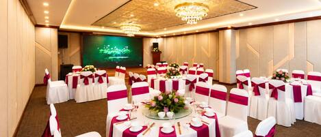 A banquet hall with round tables, white chairs with pink sashes, a large screen, and chandeliers.