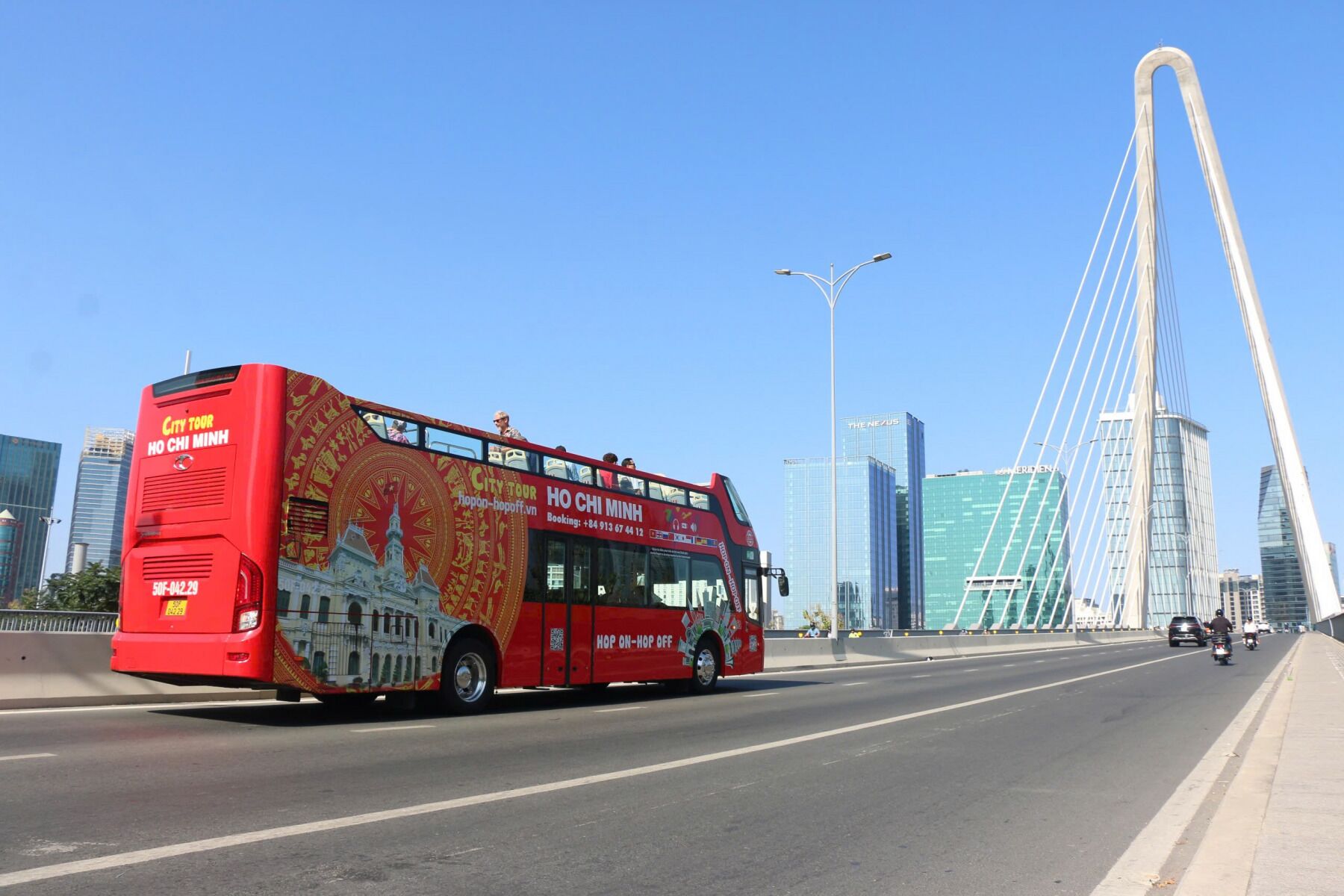 Un autobús rojo de dos pisos con la leyenda 'City Tour Ho Chi Minh' en la carrocería, circulando sobre un puente con edificios modernos al fondo.