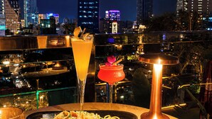 A table with a glass of orange cocktail, a red cocktail, and a plate of pasta, set against a cityscape view at night. - Happy Life Grand Hotel & Sky Bar (Ho Chi Minh City)