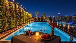 A poolside dining area with a table set for two, overlooking a cityscape at night. - Happy Life Grand Hotel & Sky Bar (Ho Chi Minh City)