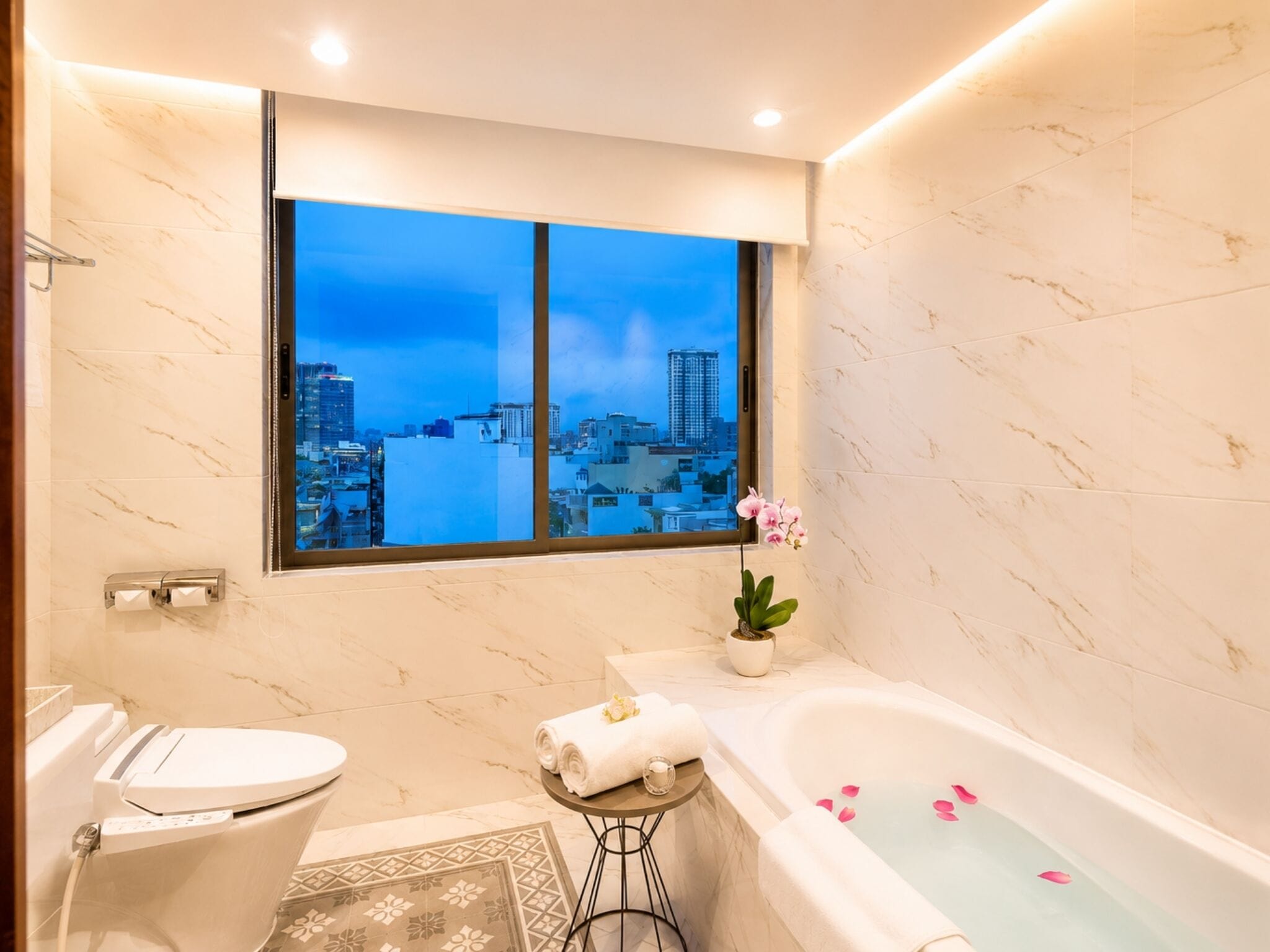 Un baño moderno con inodoro, lavamanos y bañera, con vistas a la ciudad a través de un ventanal grande.