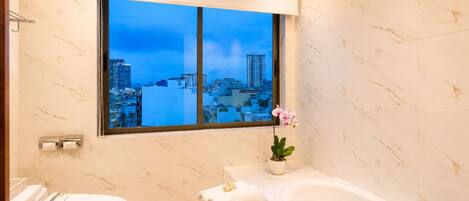 A modern bathroom with a toilet, sink, and bathtub, featuring a city view through a large window.