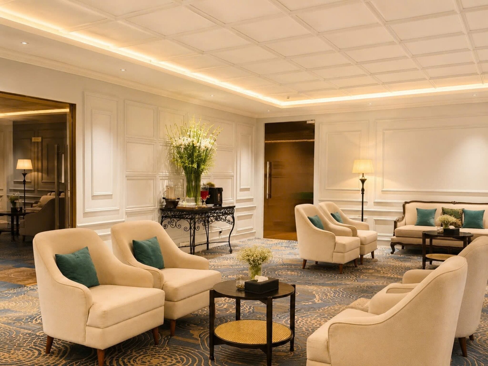 A hotel lobby with a high ceiling, a central seating area with armchairs and sofas, a patterned carpet, and a decorative vase with flowers.