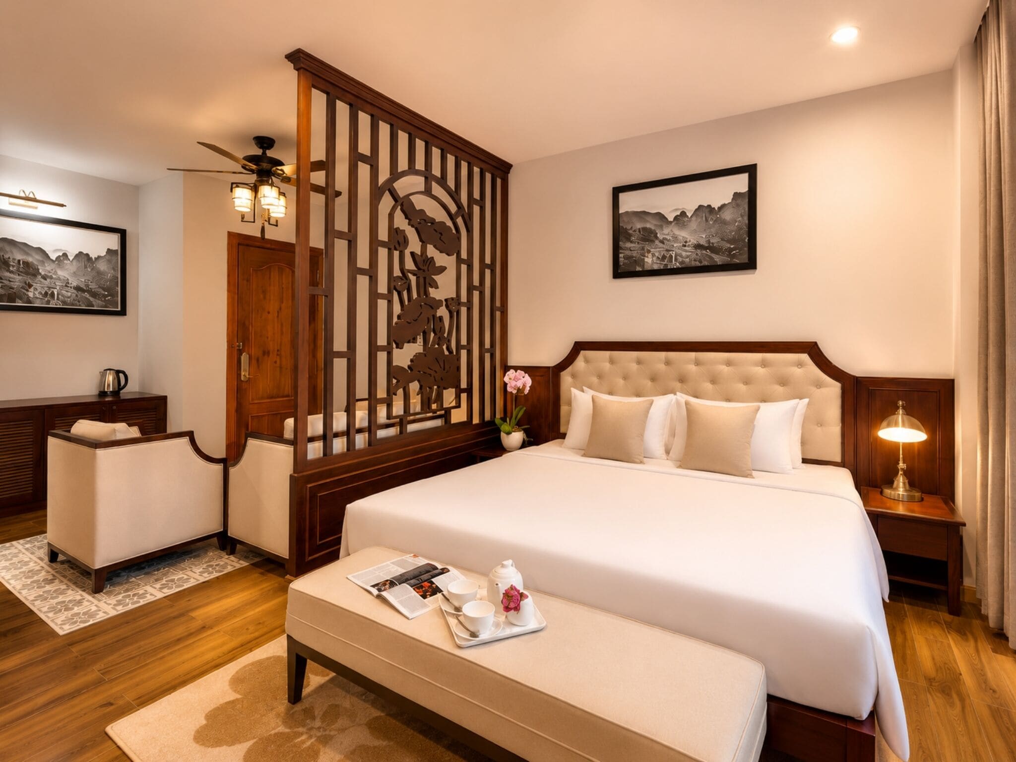 A hotel room with a large bed, a wooden partition with floral designs, a bedside table with a lamp, a small table with a tray of items, and a framed picture on the wall.