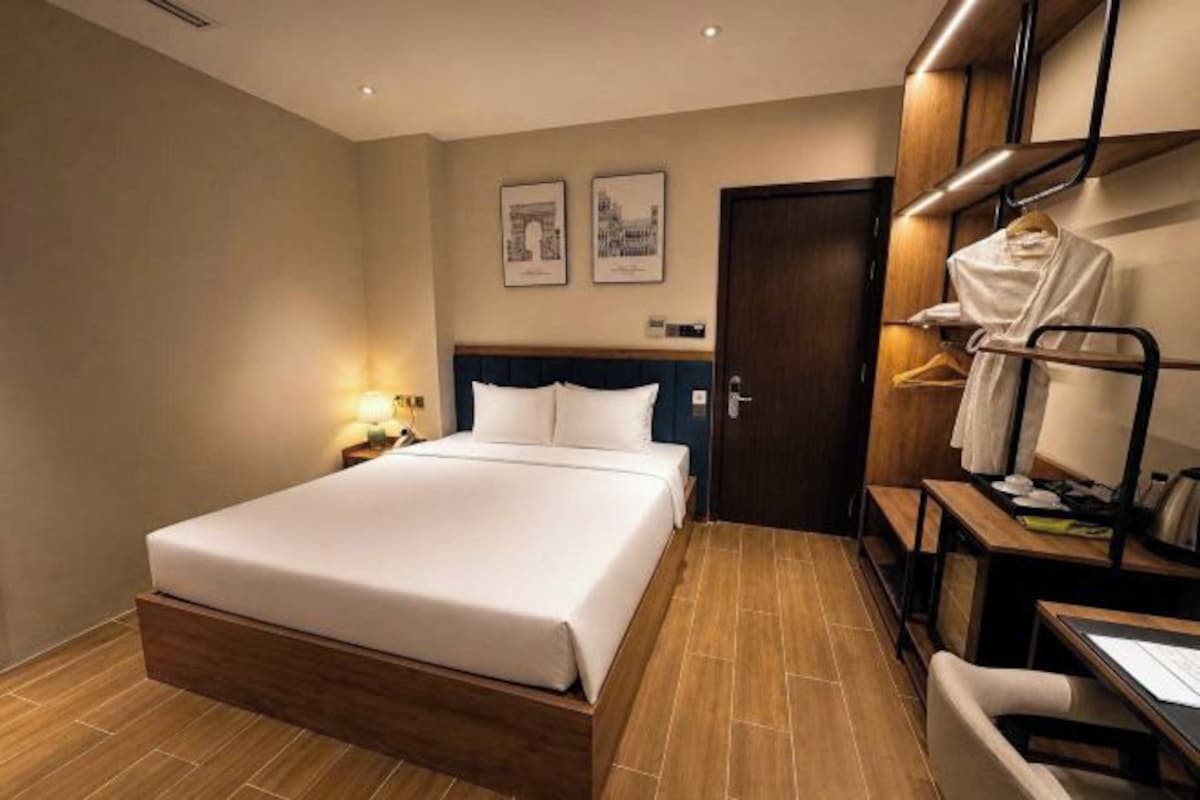 A hotel room with a bed, bedside table, lamp, and a coat rack with hanging clothes.