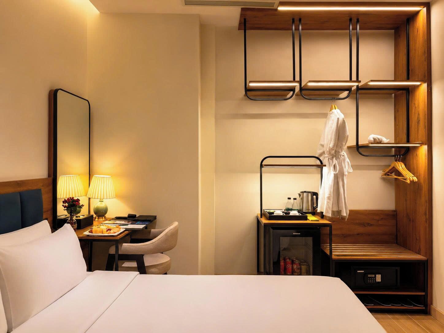 A hotel room with a bed, desk, chair, bedside table, and a wardrobe with a hanging robe.