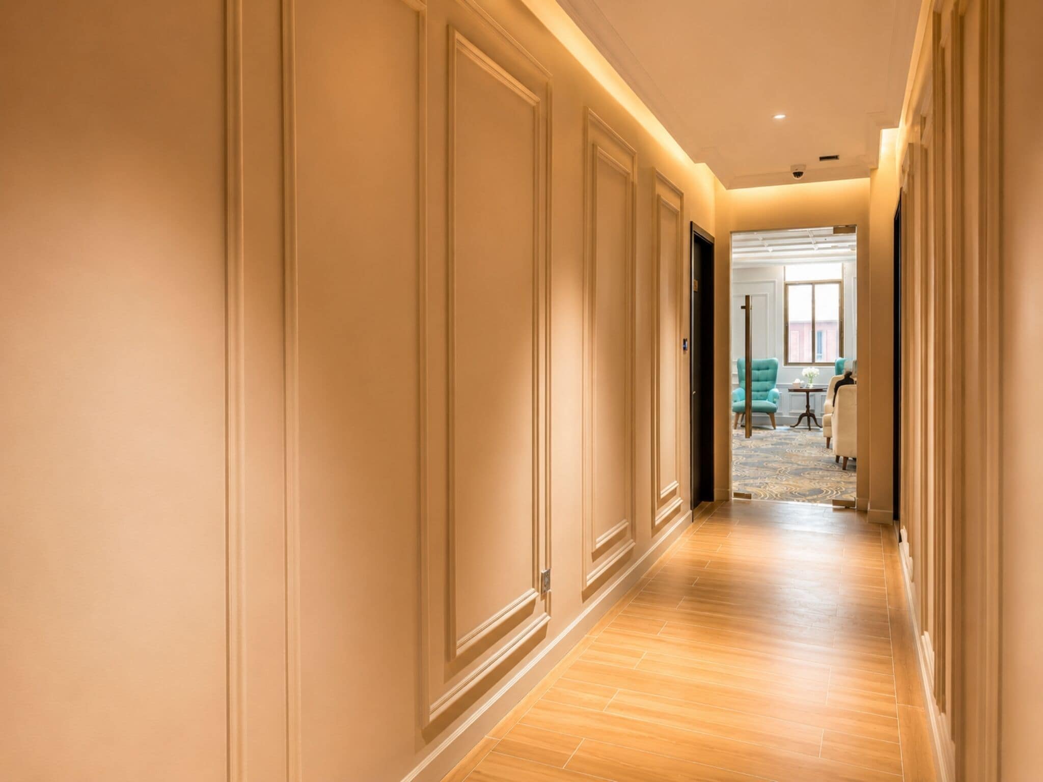 A corridor with wooden flooring and cream-colored walls, leading to a room with a blue chair and a small table.