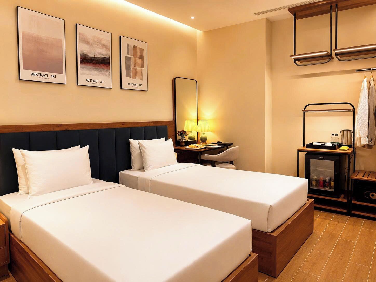 Ein Hotelzimmer mit zwei Betten, einem Schreibtisch mit Lampe, einem kleinen Kühlschrank und abstrakter Kunst an der Wand.