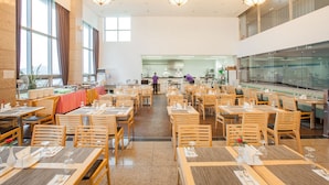Un grand espace à manger avec plusieurs tables et chaises, une cuisine à l’arrière-plan et de grandes fenêtres laissant entrer la lumière naturelle.
