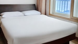Un lit bien fait, avec du linge de lit blanc, une tête de lit en bois et une vue sur la mer par une fenêtre à cadre en bois.
