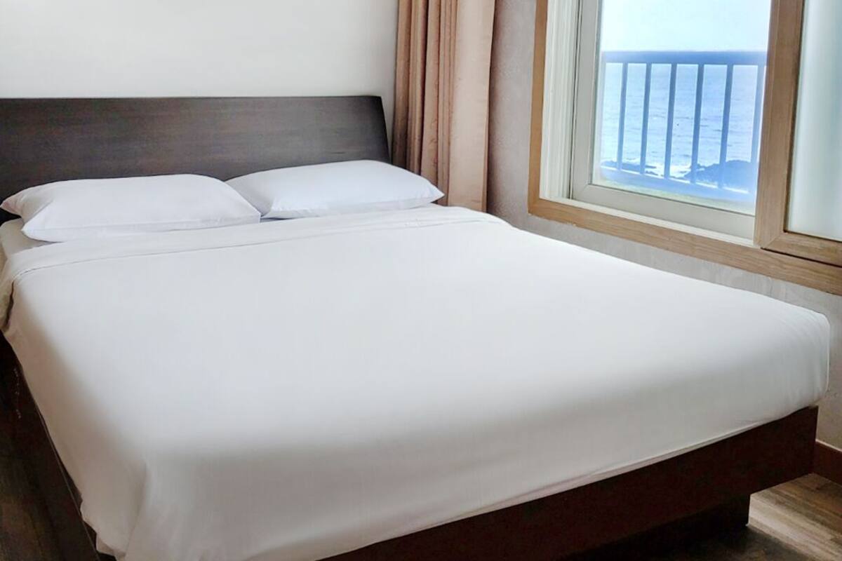 Un lit bien fait, avec du linge de lit blanc, une tête de lit en bois et une vue sur la mer par une fenêtre à cadre en bois.