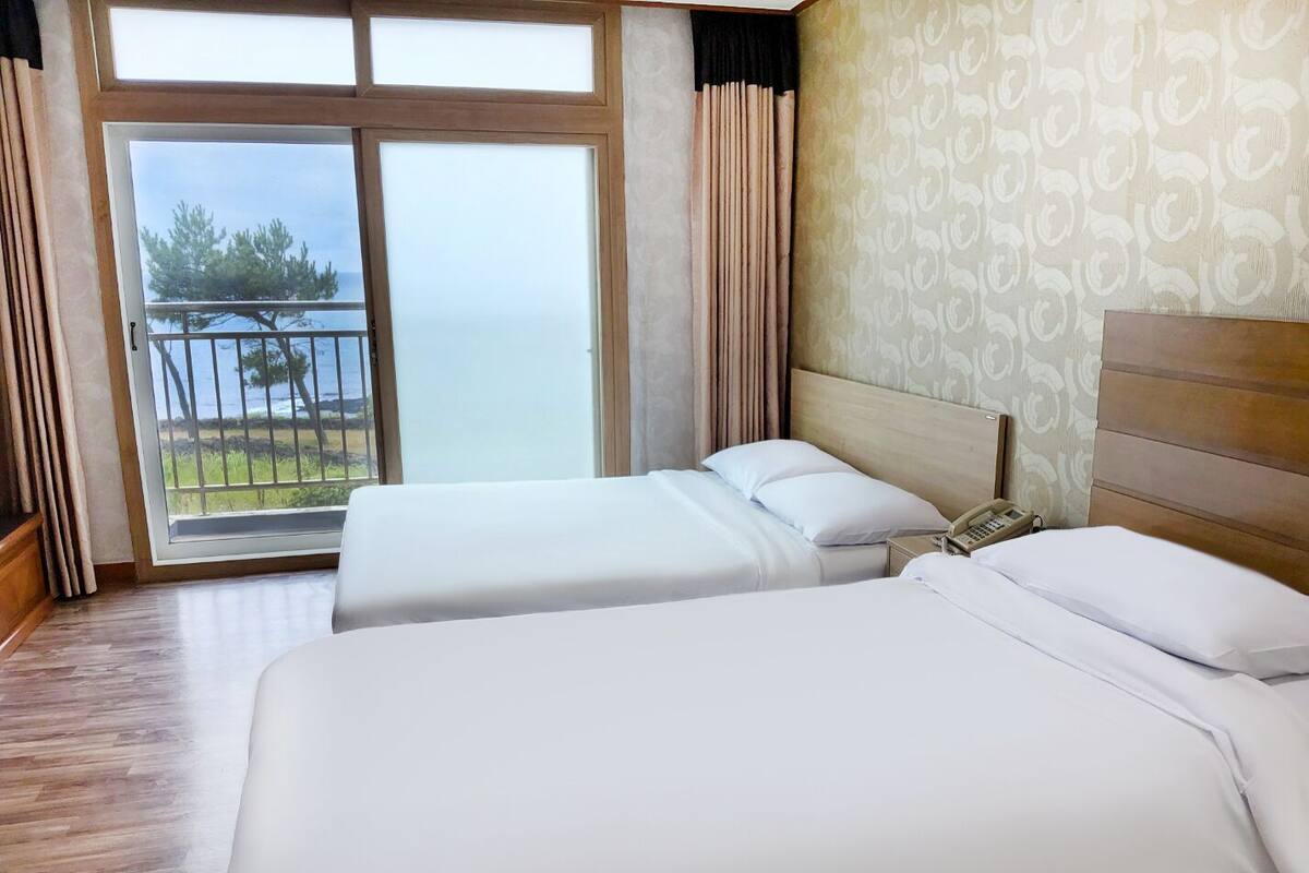Une chambre d’hôtel avec deux lits, un téléphone et une vue sur la mer, visible à travers une grande fenêtre.