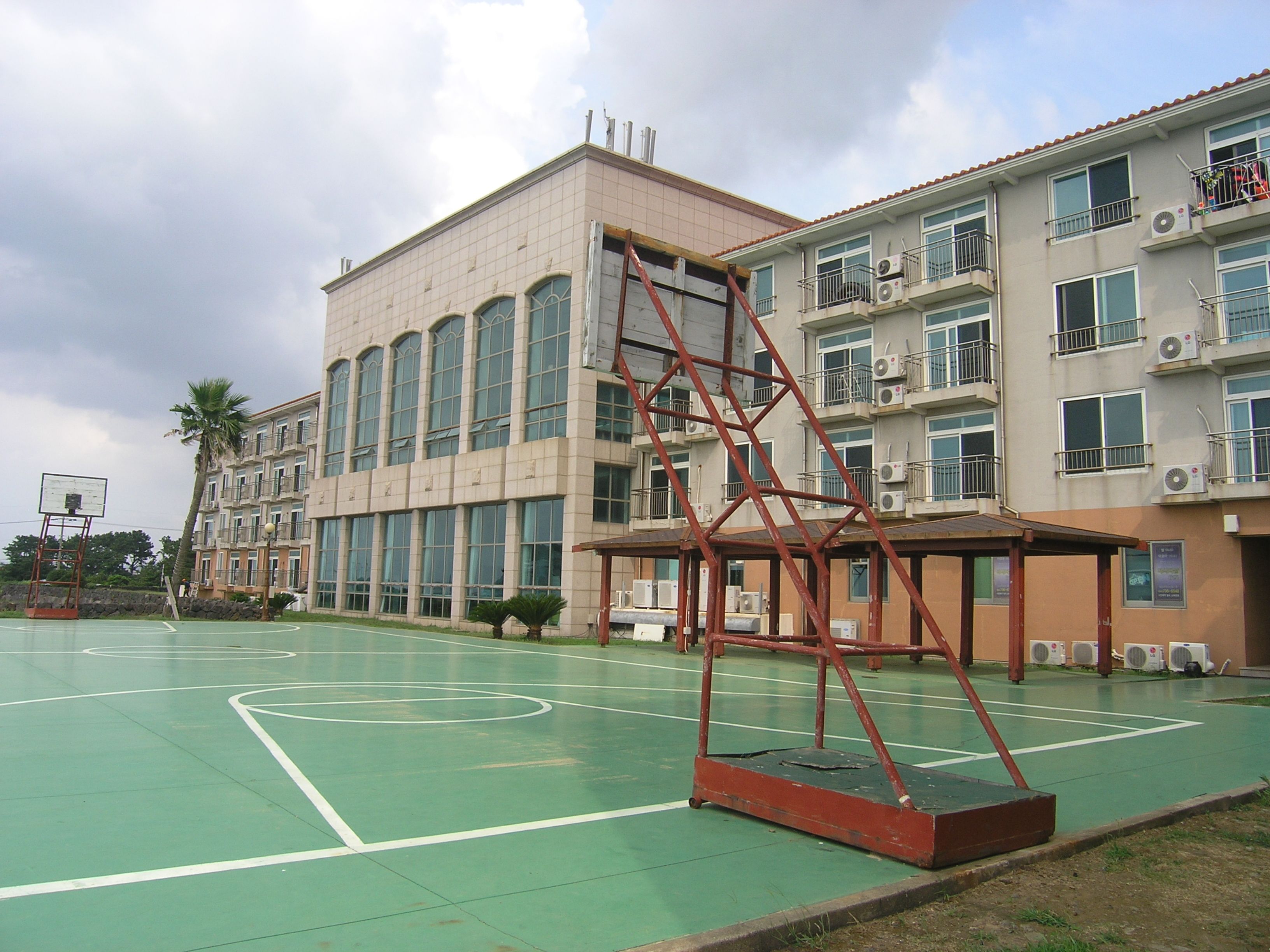A basketball court with a metal structure in the middle, surrounded by multi-story buildings with balconies and air conditioning units.