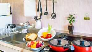 Una cocina con un lavamanos, una estufa, una olla roja, un colador con verduras, un cuenco de fruta y una planta en maceta.