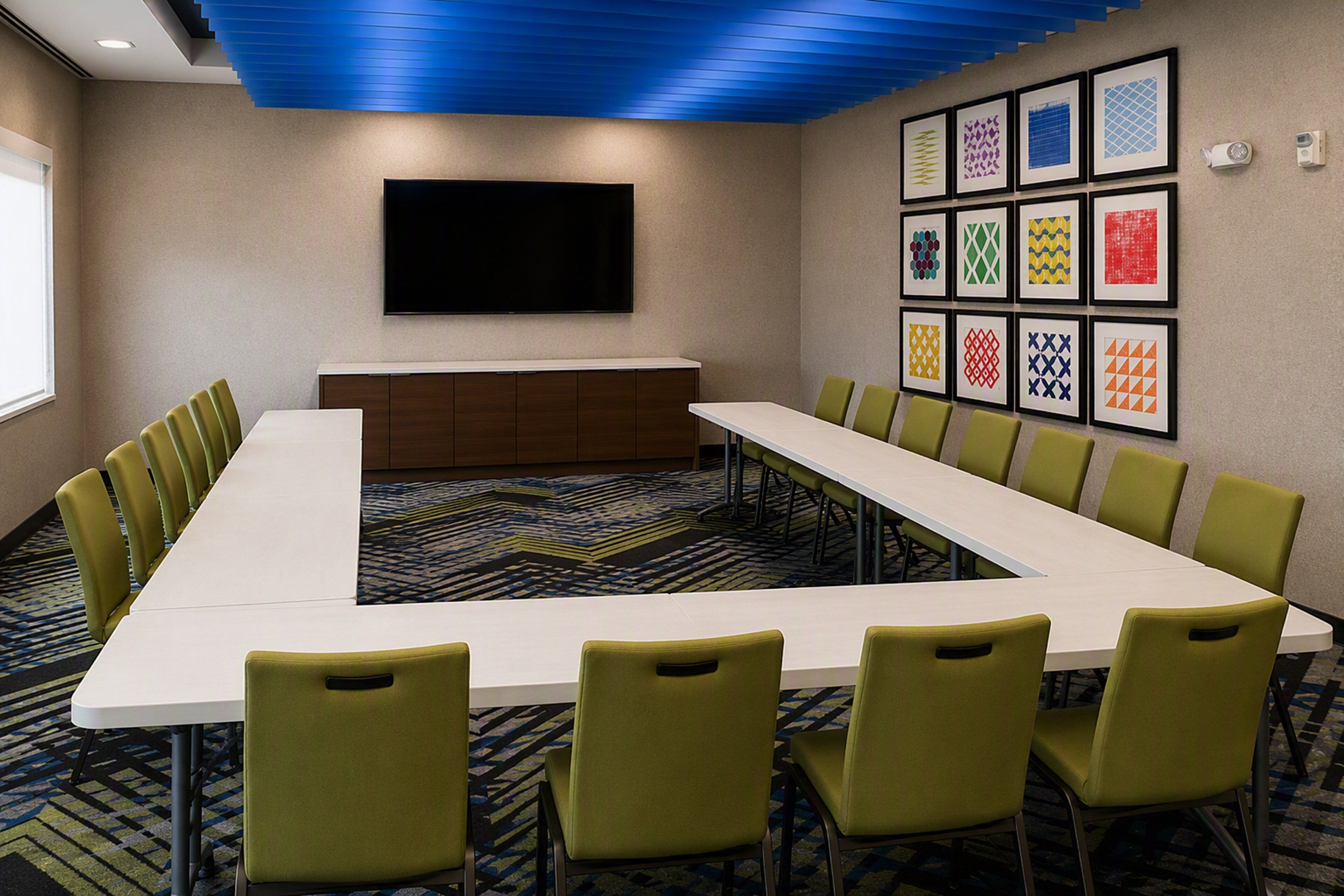 A conference room with a long table, green chairs, a flat-screen TV, and patterned wall art.