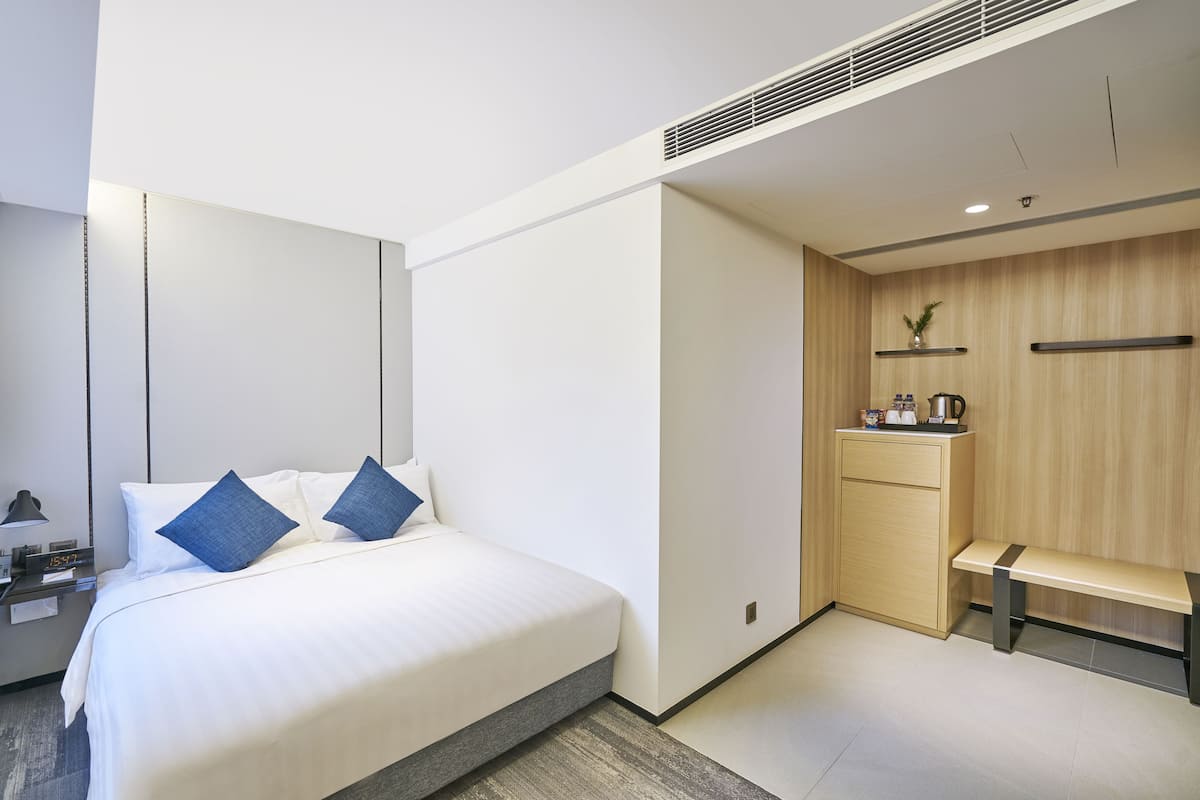 A modern hotel room with a large bed, bedside table, and a small kitchenette area.