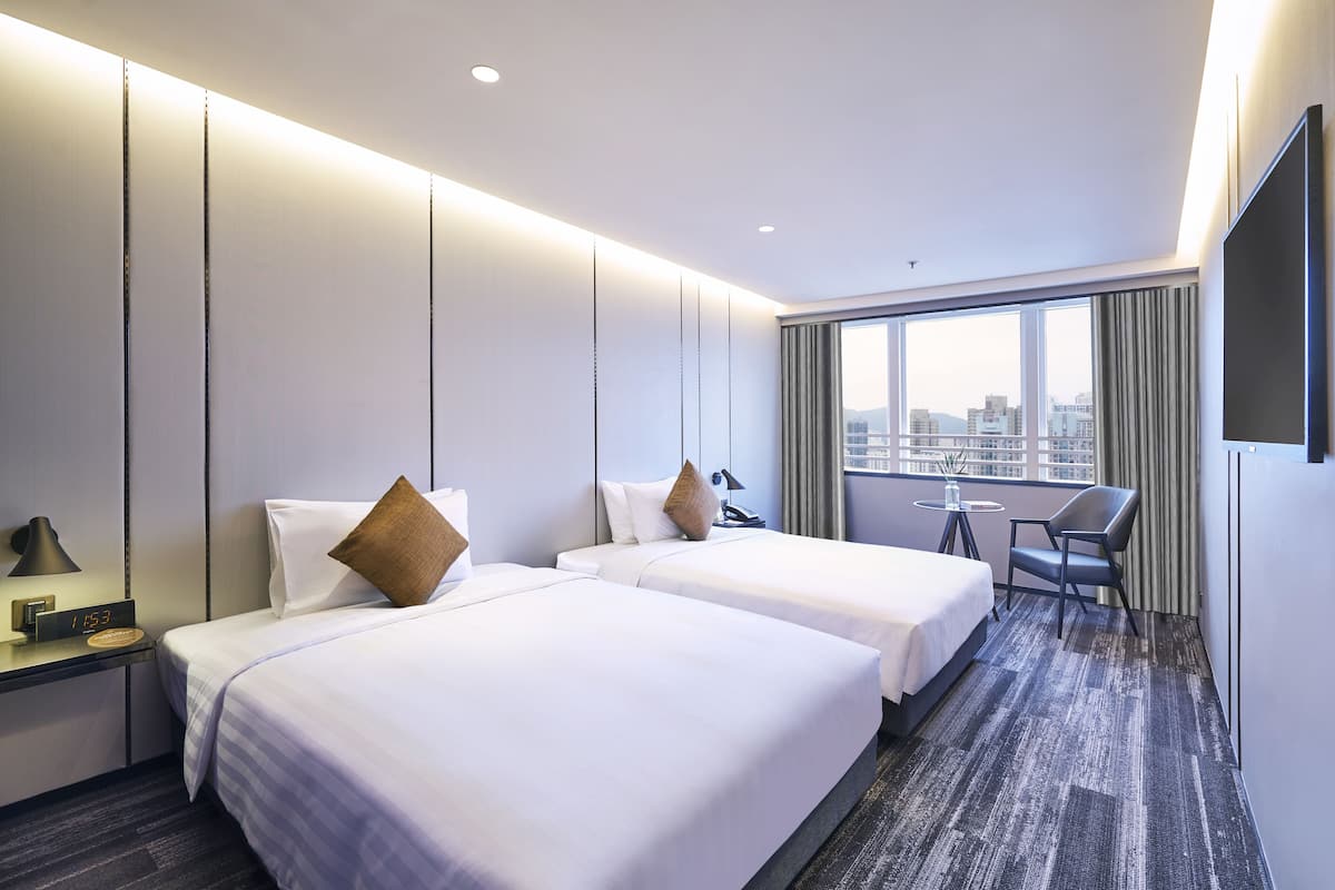 A hotel room with two beds, a desk with a lamp, a chair, and a large window with a city view.