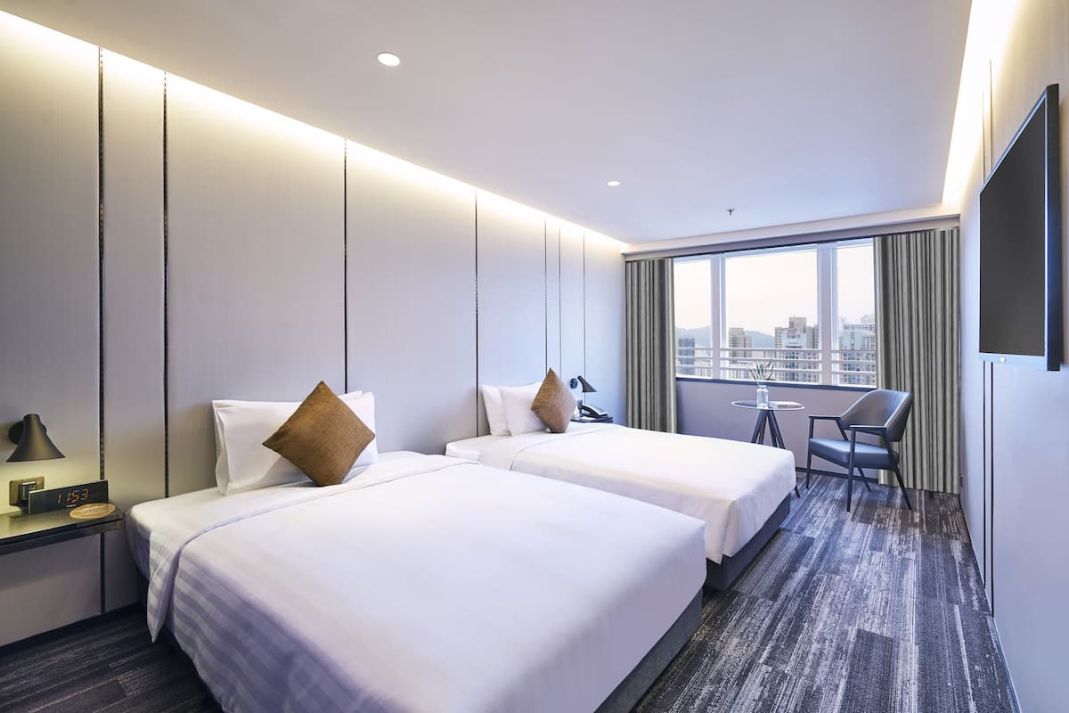 A hotel room with two beds, a desk, a chair, a TV, and a large window with a city view.