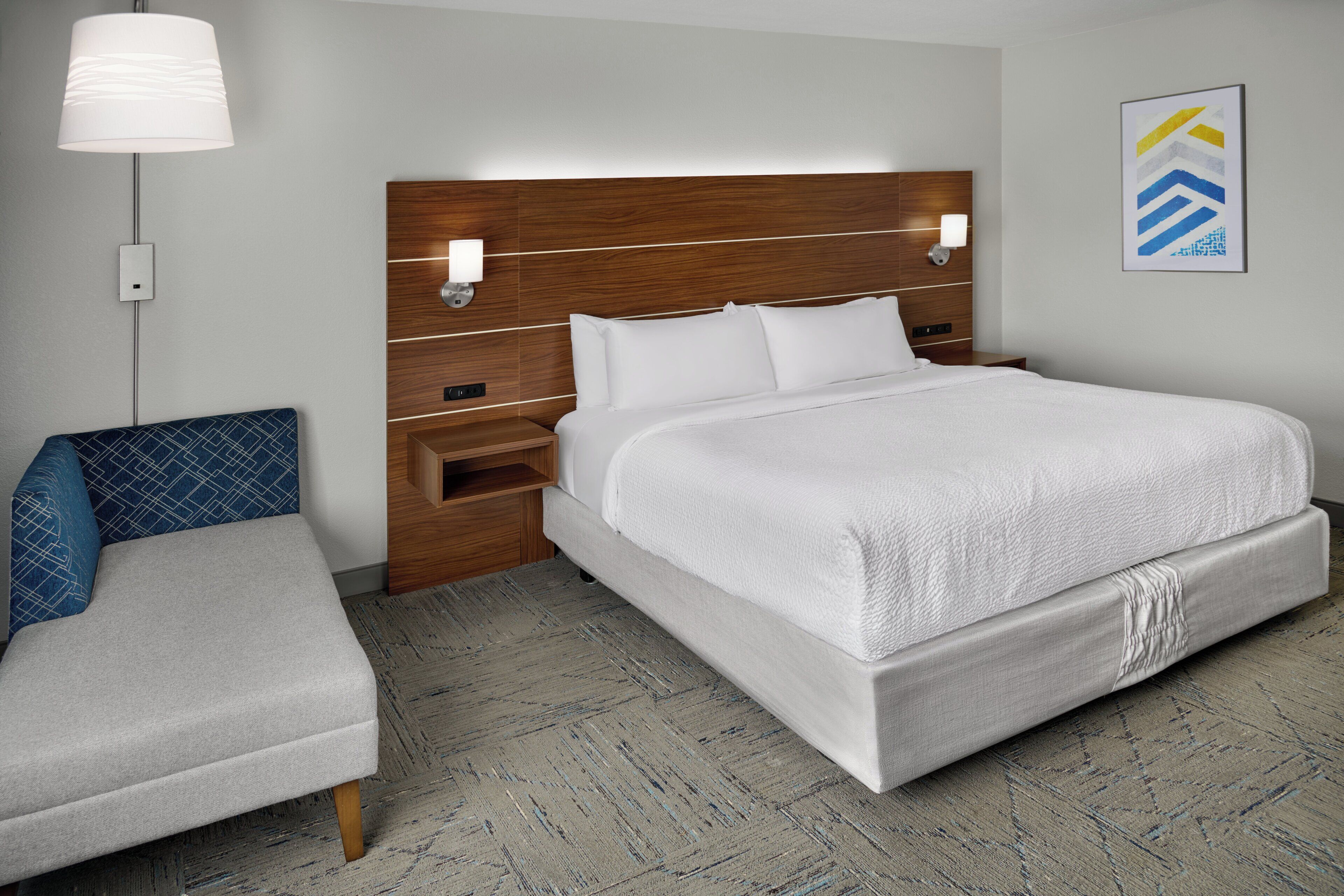 A hotel room with a large bed, a bedside table with a lamp, a chair, and a wall-mounted artwork.
