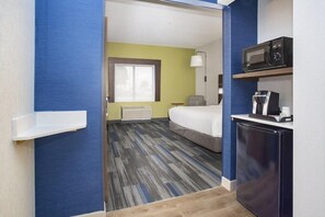 Habitación de hotel pequeña con cama, microondas, refrigerador, mesita con cafetera y ventana con persianas.
