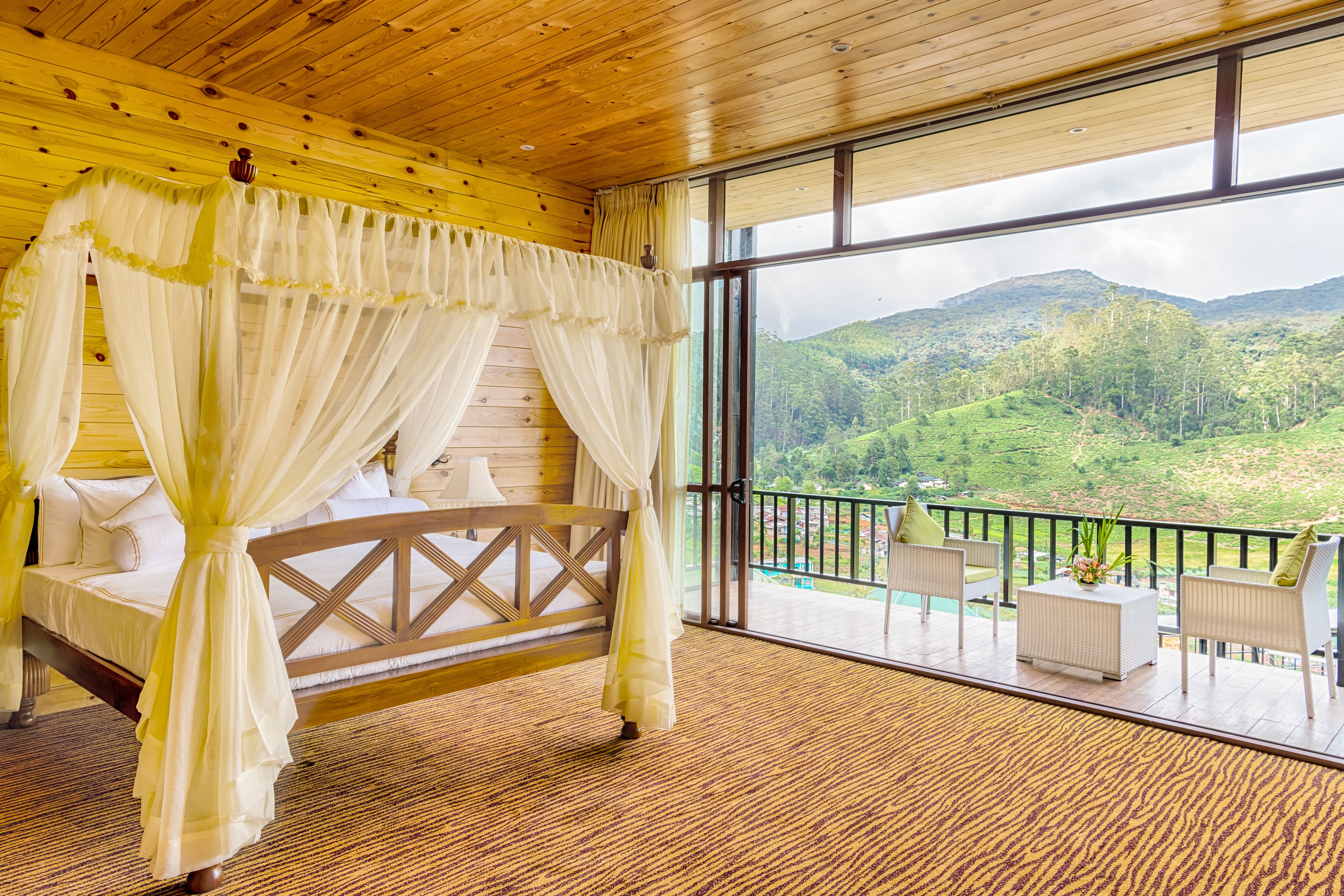 A room with a bed, a balcony with outdoor furniture, and a view of a green landscape.