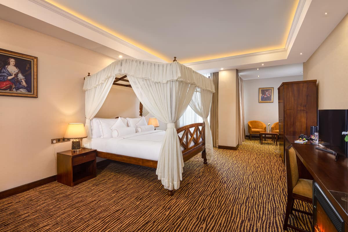 A hotel room with a four-poster bed, a desk with a flat-screen TV, and a seating area with two chairs.