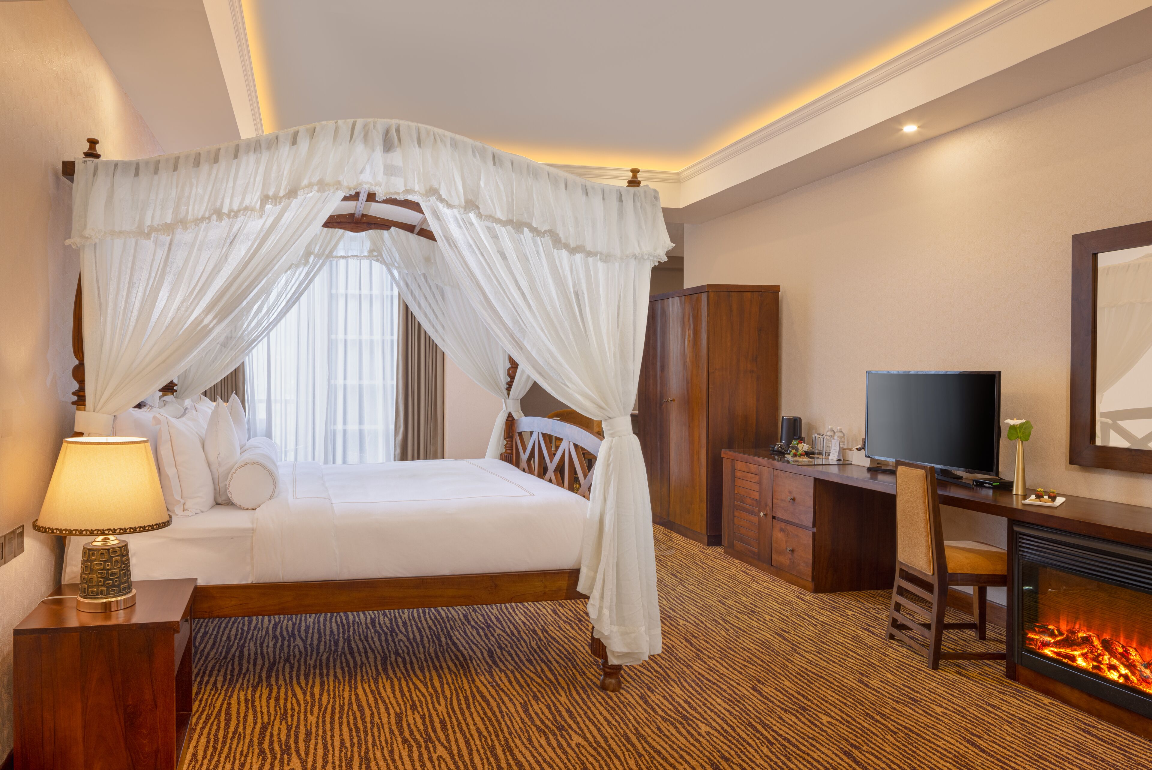 A hotel room with a four-poster bed, a desk with a flat-screen TV, a fireplace, and a large window with curtains.