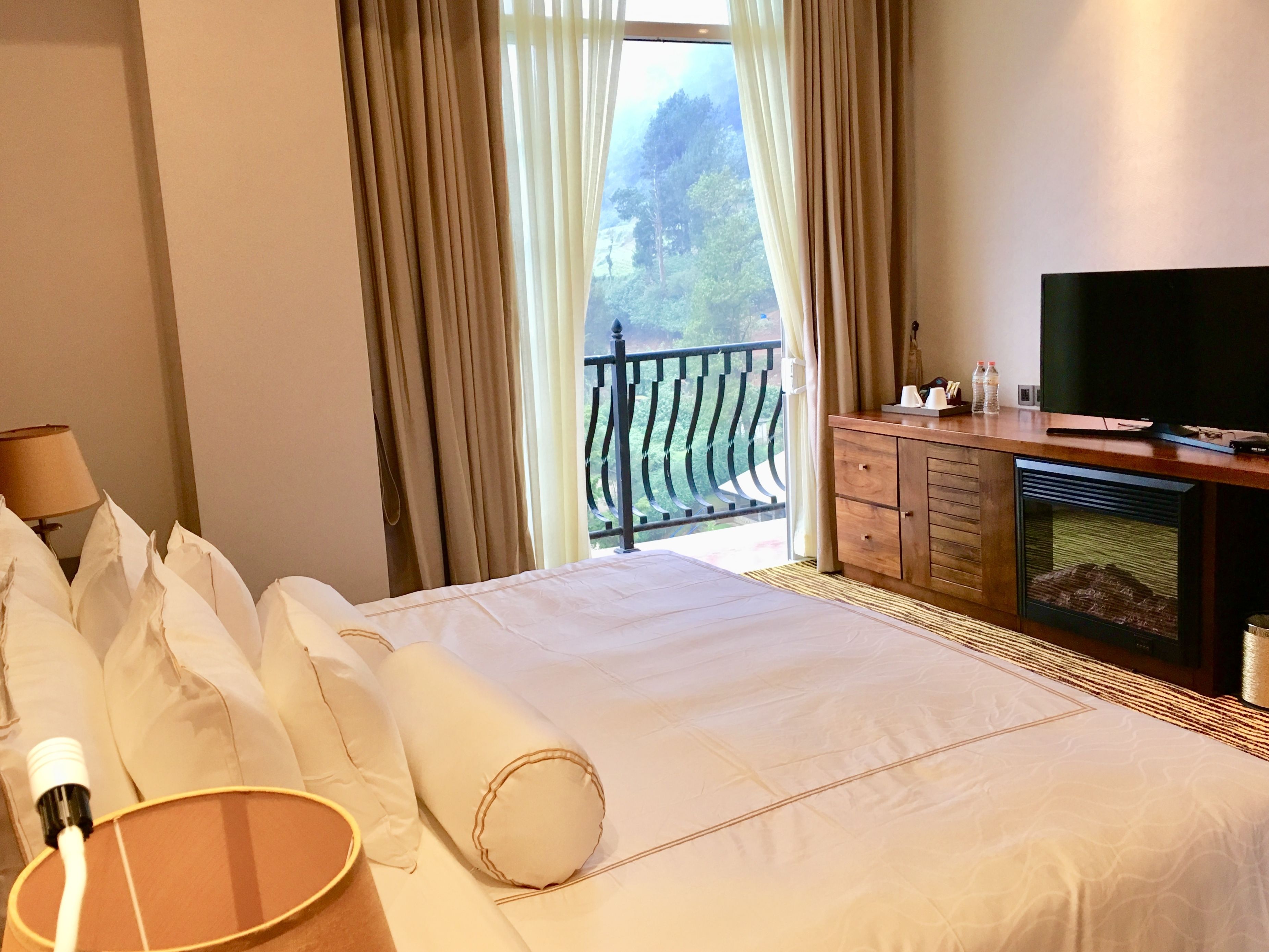 A hotel room with a large bed, a television, a wooden dresser, and a balcony with a view of trees.